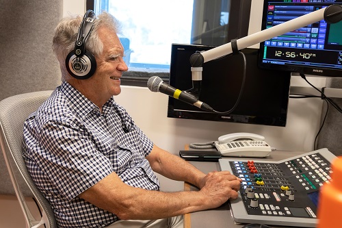 A man sits in front of microphone in a radio studio with his hands near the control panel