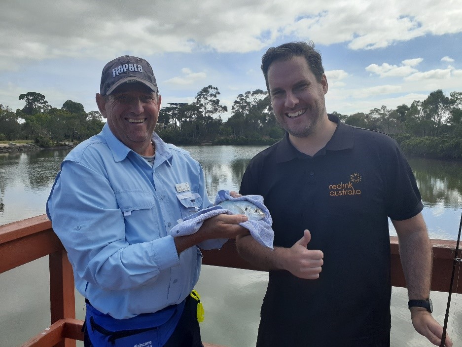 "Alan (left) holding a fish he recently caught while standing on a dock with a Fish Care instructor."