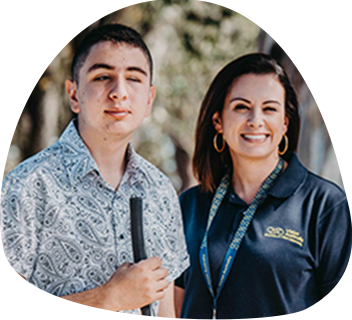 A Vision Australia staff member wearing a navy polo is standing beside a teenage boy who is holding a cane. They are both looking at the camera.