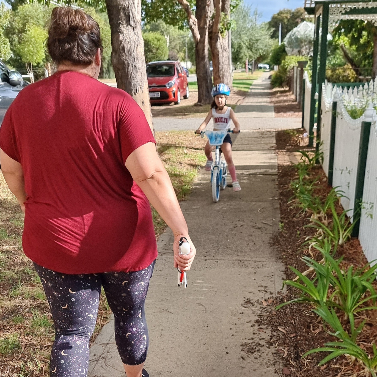 "Sarah walking down a suburban street with one of her kids on a bicycle riding towards her."