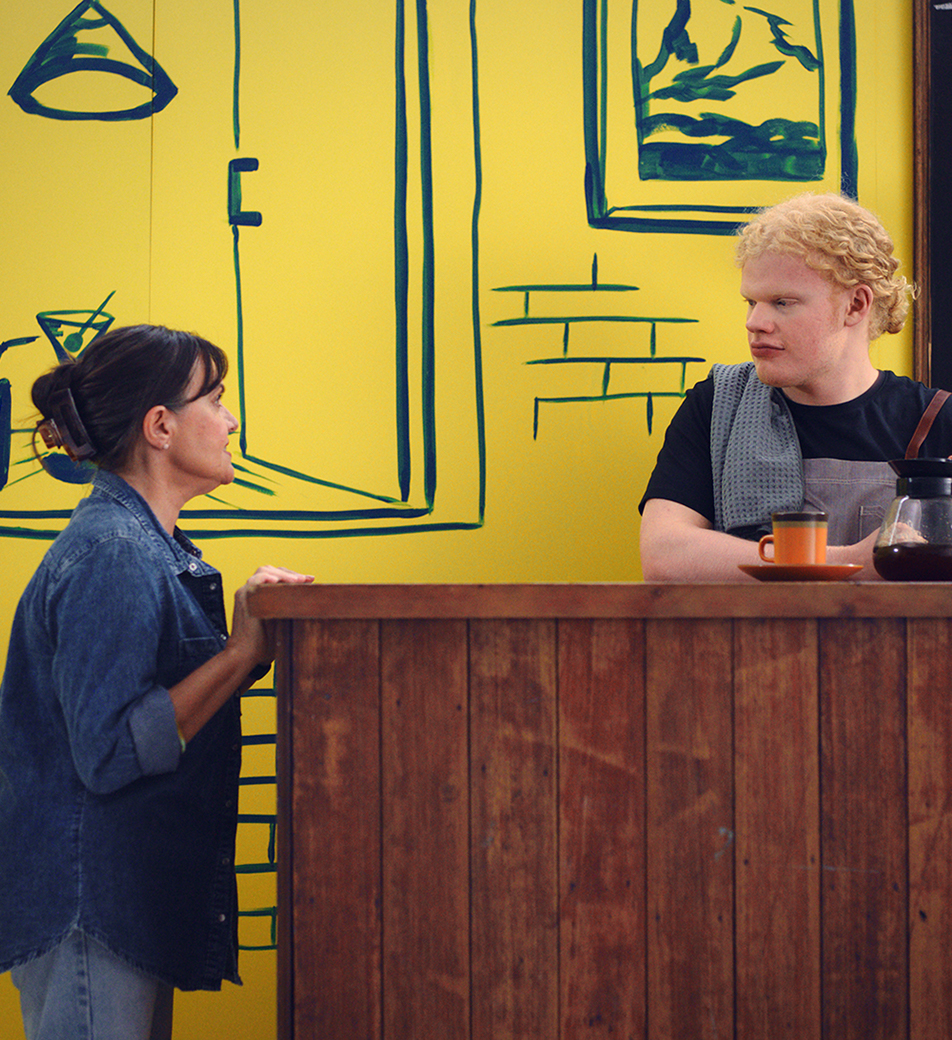 A woman wearing a denim shirt and jeans is standing at a wooden bar, speaking to a blonde man in a black t-shirt and grey apron behind the bar. There is a blackboard menu behind him with a list of 7 cheese toastie options. They are both in front of a yellow and navy painted café scene.