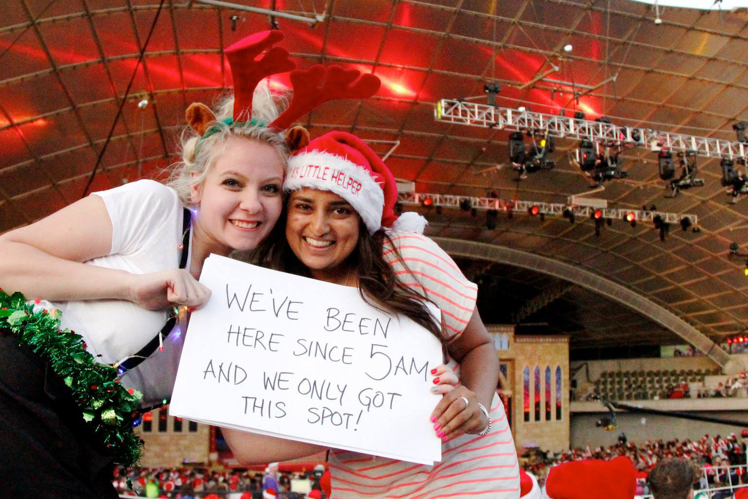 Cheery friends holding a sign in front of the Sidney Myer Music Bowl, signs reads 'We've been here since 5am and only got this spot!'