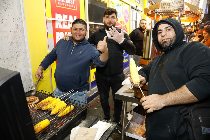 Three men pose for a photo, surrounding a hot plate cooking corn cobs