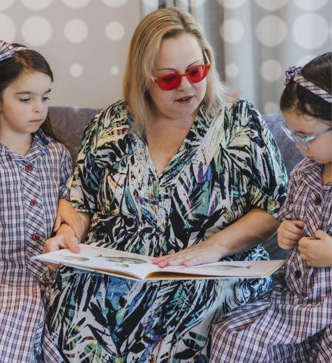 A lady is sitting between 2 little girls in school uniform, she is showing them a picture book.