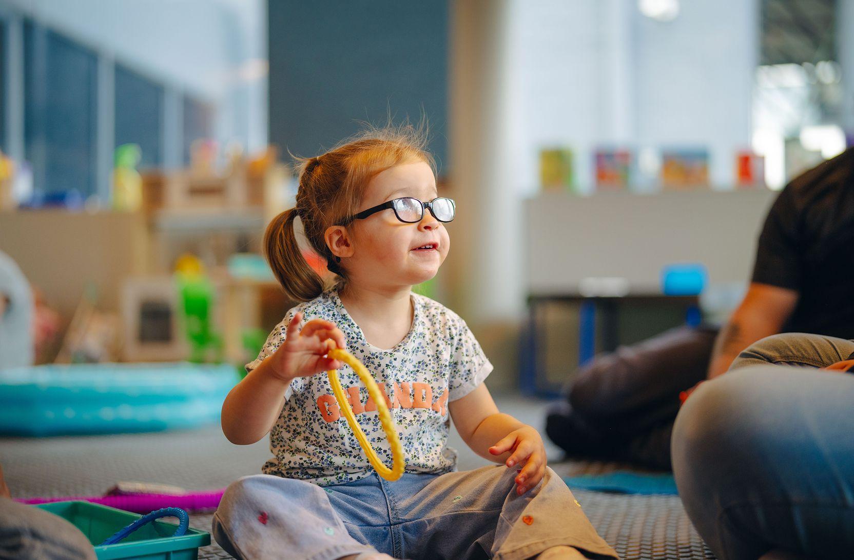A toddler girl is sitting on the ground, playing with toys, an adult sitting in the foreground.