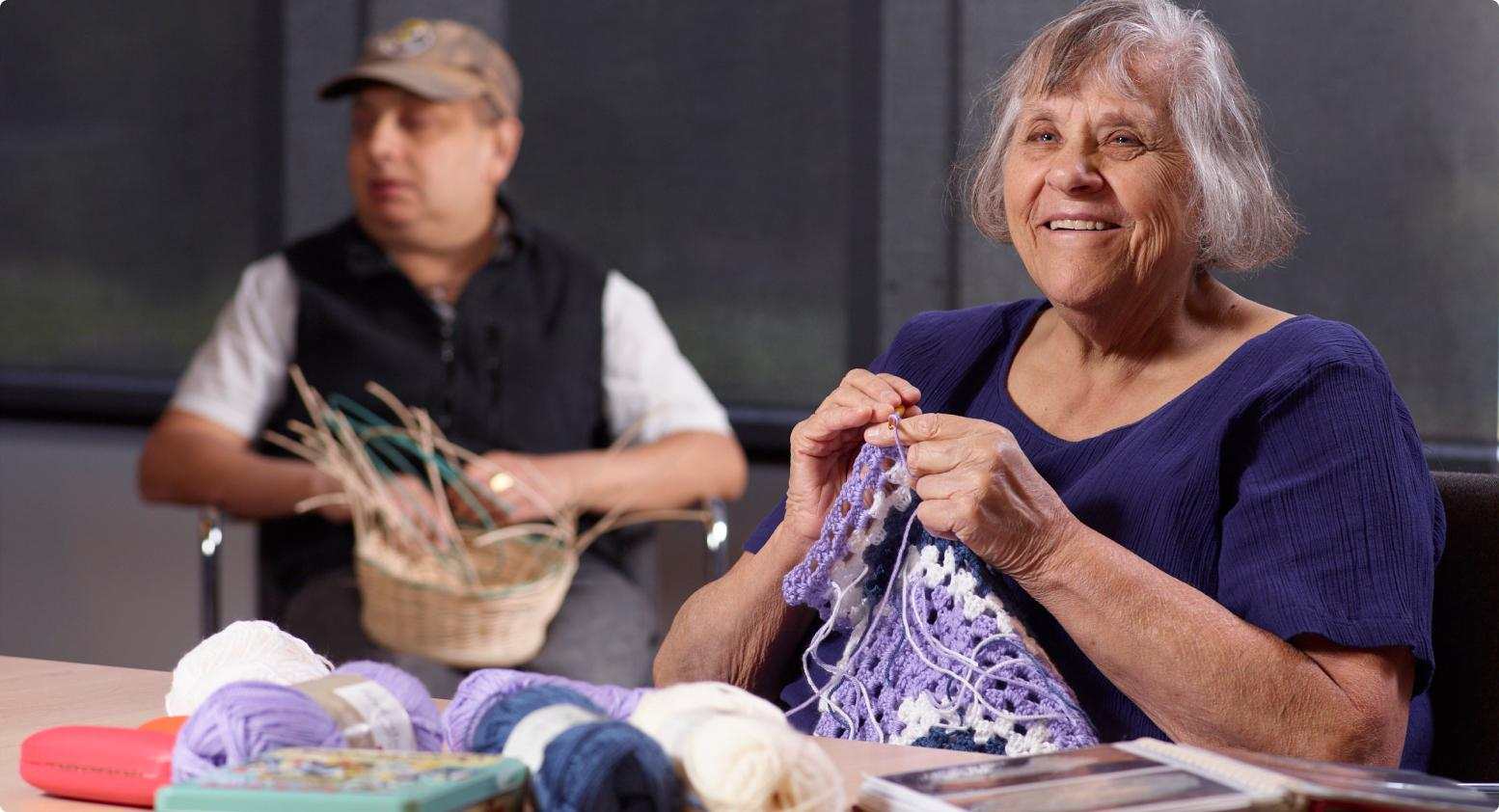 A woman smiling as she knits, seated at a table with various yarns in front of her. In the background, a man is focused on weaving a basket.