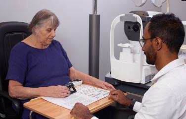 A woman in a purple T-shirt uses a magnifying glass while speaking with her eye health professional in a lab coat in his office.