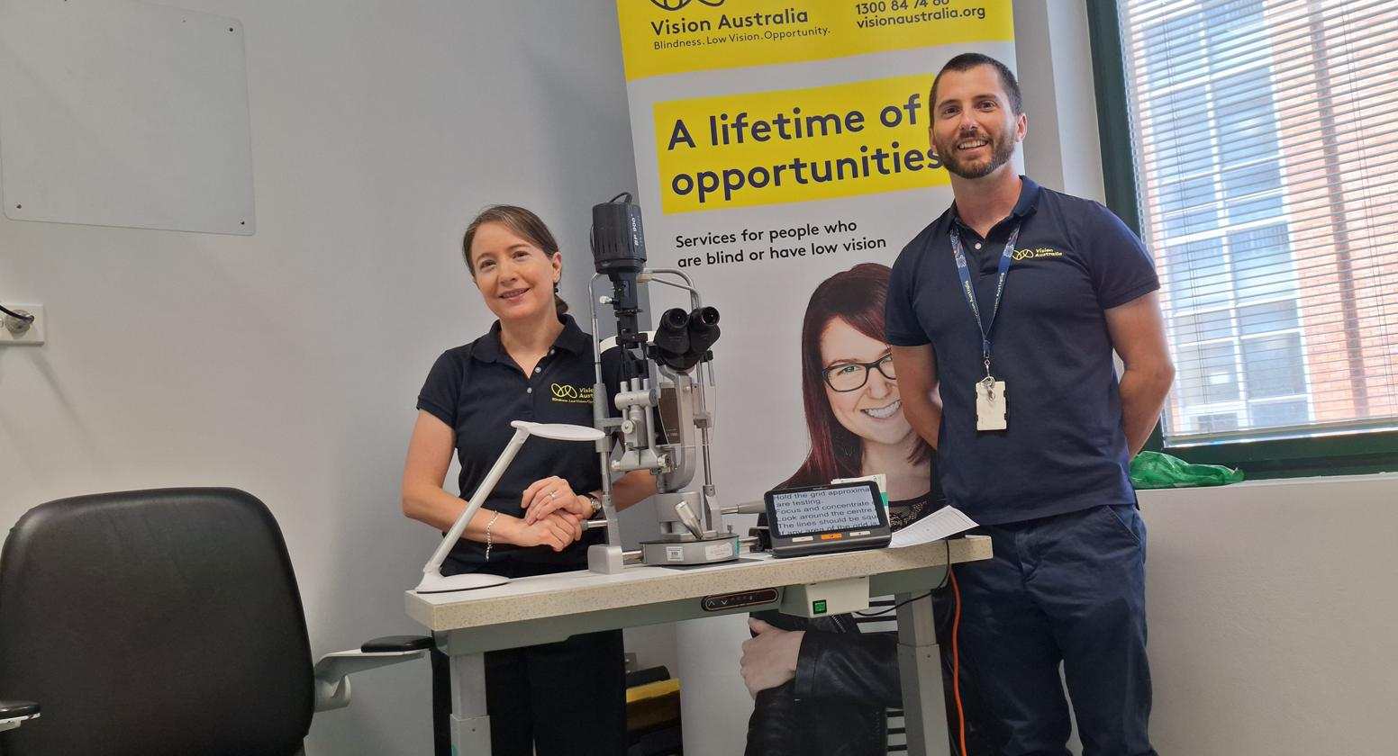 A Vision Australia optometrist and occupational therapist are smiling and standing behind a desk with assistive technology at the Know Your Vision clinic at the Sydney Eye Hospital.