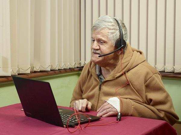An older man sitting at a laptop. He is wearing a headset and talking to someone on screen.