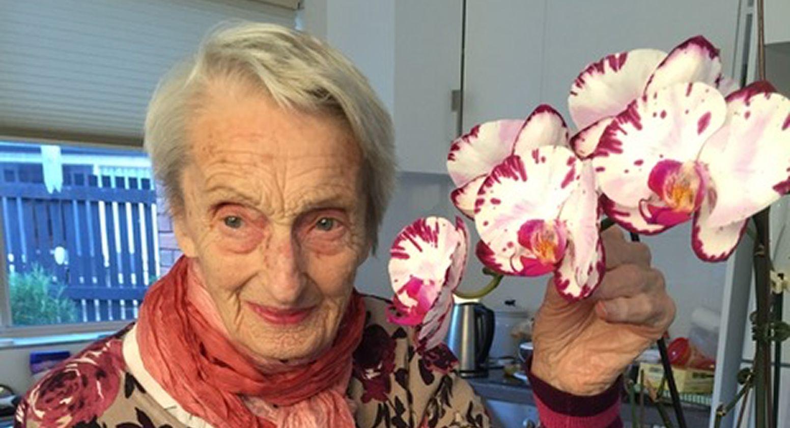 A woman is smiling, she is wearing a scarf, a floral top, she has grey hair and is holding a stem of white and pink orchid flowers.